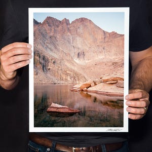 May include: A framed landscape print featuring a mountain range reflected in a calm lake. The mountains are a mix of tan and brown hues, with a clear blue sky above. The water reflects the mountains and sky, creating a symmetrical image.
