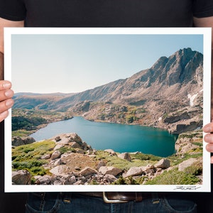 May include: A panoramic view of a turquoise lake nestled in a mountain valley. The lake is surrounded by rocky, green hills and a towering mountain peak in the background. The sky is a clear blue.