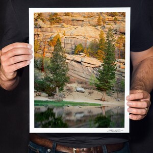 May include: A framed landscape photograph featuring a serene lake reflecting the surrounding trees and rocky cliffs. The image showcases a natural scene with a clear sky, lush greenery, and a sense of tranquility. The photograph is held by a person.
