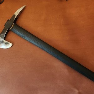 May include: A hand-forged metal war hammer featuring a curved axe blade on one side and a pointed pick on the opposite side. The tool has a long, straight, dark-stained wooden handle that is securely attached to the metal head with visible welding.