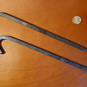 Puede incluir: Dos estacas de tienda de campaña de metal negro con un gancho en un extremo. Las estacas miden aproximadamente 30 cm de largo y están sobre una superficie marrón.