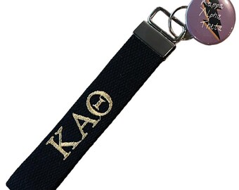 May include: Black fabric keychain with gold embroidered "KAΘ" letters. A round button with "Kappa Alpha Theta" text and a lightning bolt design is attached. The keychain has a silver metal clasp.