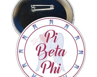 May include: A circular pin-back button featuring a white background with the text Pi Beta Phi in dark pink script. The design includes a light pink heart with an arrow and large light blue angel wings. A border of small blue wings surrounds the edge.