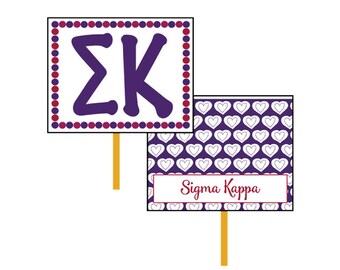 May include: Two purple and white signs with a stick attached to the bottom. The top sign has a purple "ΣΚ" on a white background with a red and white polka dot border. The bottom sign has a white heart pattern on a purple background with a white rectangle at the bottom that says "Sigma Kappa" in red.