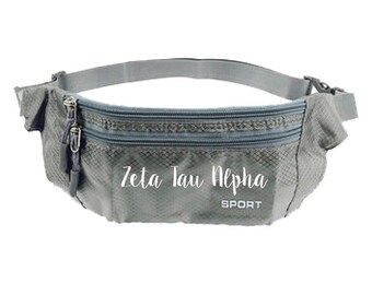 May include: Gray fanny pack with two zippered compartments and an adjustable strap. The text "Zeta Tau Alpha SPORT" is printed on the front. The bag is made of a lightweight, textured material.