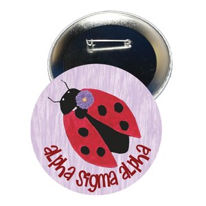 May include: A round button pin featuring a red ladybug with black spots and a purple flower on a light purple background. The words "alpha sigma alpha" are written in red around the ladybug. The pin has a silver metal backing.