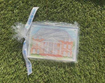 May include: A rectangular clear plastic package containing a printed illustration of a multi-story red brick building with white columns. A white ribbon with the blue Greek letters ΑΔΠ is tied around the top corner of the plastic packaging.