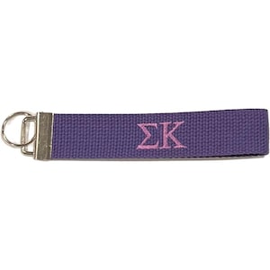May include: A purple key fob with a silver metal clasp and ring. The key fob has the embroidered letters ΣΚ in pink. The key fob is made of a woven fabric.