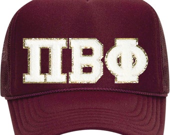 May include: A maroon trucker hat with a mesh back and a curved brim. The front of the hat features the Greek letters ΠΒΦ in white with gold glitter trim. The hat has a maroon rope detail above the brim.