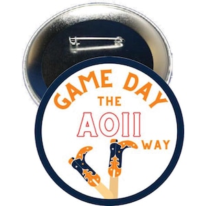 May include: A circular pin-back button with a white background and a dark blue border. The text reads GAME DAY THE AOII WAY in orange and red. Two orange and dark blue cowboy boots with star accents are illustrated at the bottom of the button.