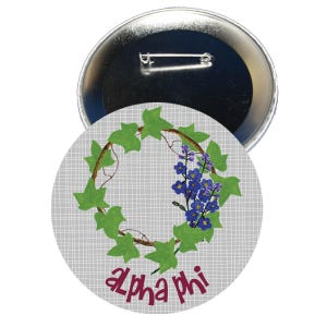 May include: A circular badge with a silver metal back and a floral design. The design features a wreath of green leaves and blue flowers around a brown vine. The text "Alpha Phi" is in maroon script.