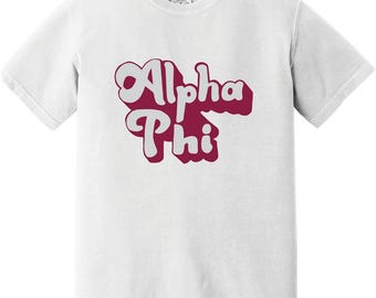 May include: A white short-sleeve t-shirt featuring the text Alpha Phi printed in a bold, retro-style font. The lettering is light pink with a dark maroon drop shadow, creating a layered effect across the center of the chest area of the garment.