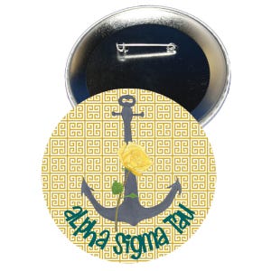 May include: A circular badge with a gold and teal design. It features a dark grey anchor with a yellow rose. The words "alpha sigma tau" are written in teal. The background has a gold Greek key pattern.
