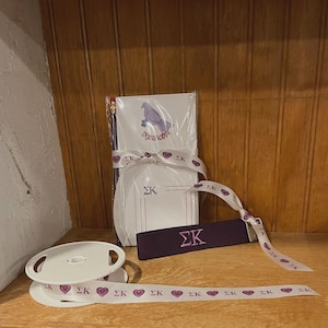 May include: A collection of Sigma Kappa sorority merchandise, including a notepad with a dove graphic, pencils, a ribbon, and a purple band. The ribbon and band feature the letters "ΣΚ". The ribbon also has heart graphics.