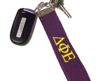 May include: A purple key fob with yellow embroidered Greek letters, attached to keys and a Jeep keyless entry remote. The key fob is made of a textured fabric.