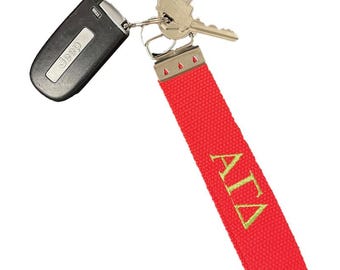 May include: A red fabric keychain with gold embroidered Greek letters, attached to keys and a black Jeep key fob. The keychain is a vibrant red color, and the Greek letters are in a gold color.