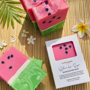 Könnte beinhalten: Handgemachte Seifenstücke in Wassermelonenform mit rosa, grünen und weißen Schichten. Die Seife hat schwarze Samendetails und Glitzerakzente. Eine Seifenbox ist ebenfalls sichtbar, mit dem Text "Watermelon Sugar HANDCRAFTED SOAP".
