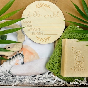 Puede incluir: Un set de regalo para beb&eacute; en una caja, que incluye un letrero de madera con "hello world" y espacio para detalles, un body con estampado de cebra, un mordedor, un pa&ntilde;o verde y una pastilla de jab&oacute;n "Baby Soap" con dise&ntilde;o de jirafa. La caja est&aacute; llena de papel.