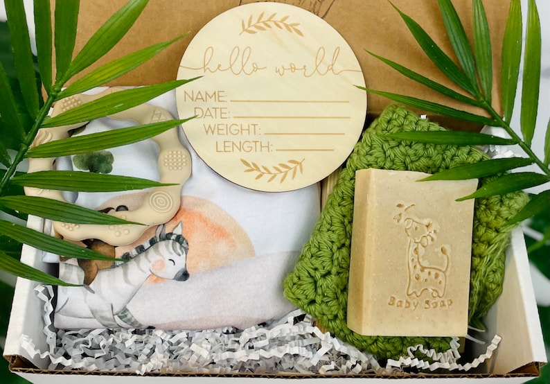 Puede incluir: Un set de regalo para beb&eacute; en una caja, que incluye un letrero de madera con "hello world" y espacio para detalles, un body con estampado de cebra, un mordedor, un pa&ntilde;o verde y una pastilla de jab&oacute;n "Baby Soap" con dise&ntilde;o de jirafa. La caja est&aacute; llena de papel.