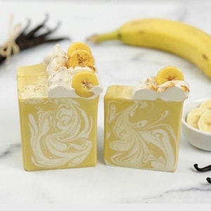 K&ouml;nnte beinhalten: Zwei quadratische Seifenst&uuml;cke in Gelb mit wei&szlig;en Wirbeln, belegt mit wei&szlig;em Schaum und Bananenscheiben. Eine ganze Banane, eine kleine Sch&uuml;ssel mit Bananenscheiben und Vanilleschoten sind ebenfalls im Bild.