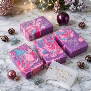 May include: Four rectangular bars of artisan soap with a swirling design in shades of pink, blue, and purple. The soap bars are arranged on a marble surface with pinecones, ornaments, and a tag that reads "Sugar Plum Dreams Artisan Soap."