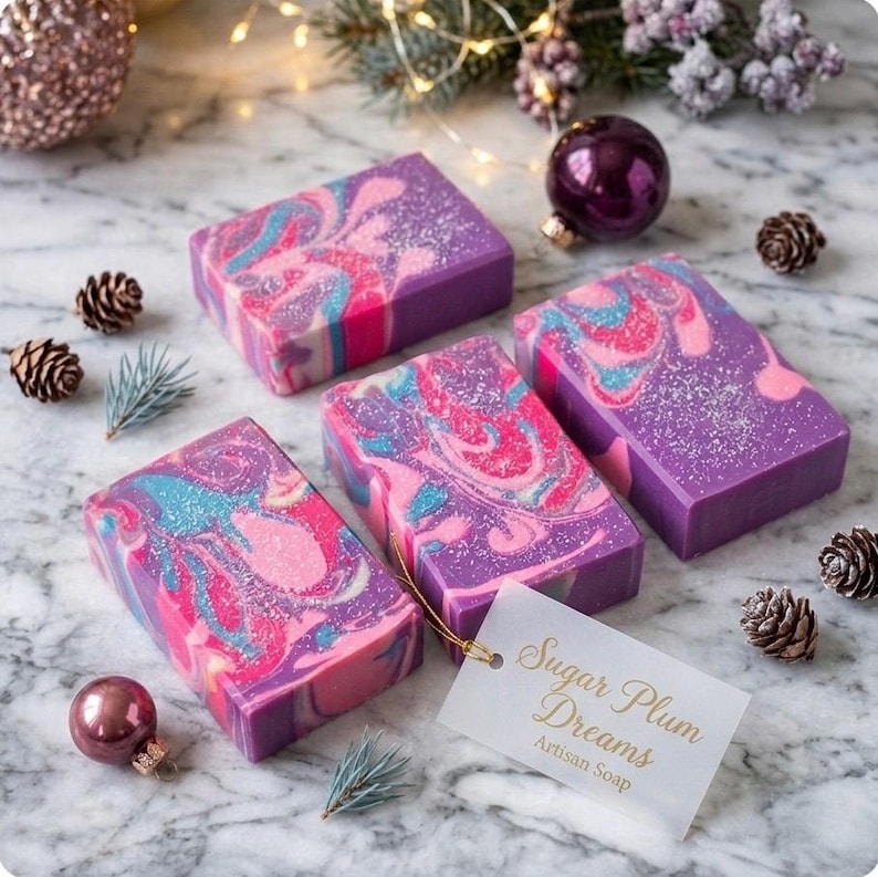 May include: Four rectangular bars of artisan soap with a swirling design in shades of pink, blue, and purple. The soap bars are arranged on a marble surface with pinecones, ornaments, and a tag that reads "Sugar Plum Dreams Artisan Soap."