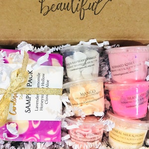 May include: A gift box filled with a variety of bath and body products. The box contains a bar of soap, a small jar of whipped body butter, and several small containers of foaming face and body scrubs. The products are all in shades of pink, purple, and white. The box is decorated with a gold ribbon and a tag that reads "Sample Pack".
