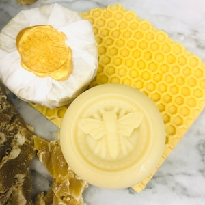 May include: Three bars of soap with a honeycomb design. The soap on the left is wrapped in white paper with a gold bee wax seal. The soap in the center has a bee design embossed on the top. The soap on the right is a honeycomb shaped bar.