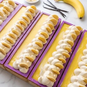 K&ouml;nnte beinhalten: Handgemachte Seifen in lila Formen, verziert mit wei&szlig;en Wirbeln und Bananenscheiben. Eine gelbe Grundfarbe ist sichtbar. Eine reife Banane, Vanilleschoten und geschnittene Bananen sind ebenfalls vorhanden.