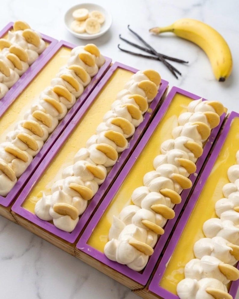 K&ouml;nnte beinhalten: Handgemachte Seifen in lila Formen, verziert mit wei&szlig;en Wirbeln und Bananenscheiben. Eine gelbe Grundfarbe ist sichtbar. Eine reife Banane, Vanilleschoten und geschnittene Bananen sind ebenfalls vorhanden.