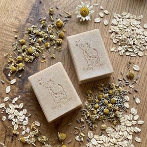Puede incluir: Dos jabones para beb&eacute;s beige con un dise&ntilde;o de jirafa y la inscripci&oacute;n "Baby Soap" est&aacute;n rodeados de flores de manzanilla, avena y capullos de lavanda sobre una superficie de madera. Tambi&eacute;n hay una margarita.