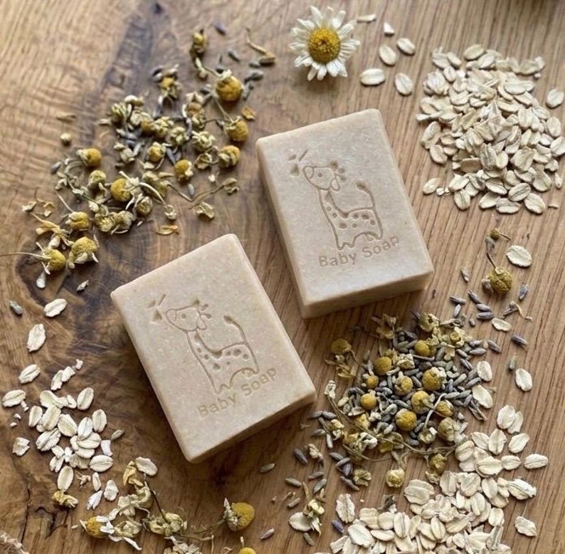 Puede incluir: Dos jabones para beb&eacute;s beige con un dise&ntilde;o de jirafa y la inscripci&oacute;n "Baby Soap" est&aacute;n rodeados de flores de manzanilla, avena y capullos de lavanda sobre una superficie de madera. Tambi&eacute;n hay una margarita.