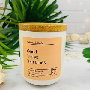 May include: A white candle with a bamboo lid. The label reads "Carolina Organics Soap Co. Good Times, Tan Lines" and "She had dumps like truck, truck, truck. Soy Wax Candle 8oz/227gr".