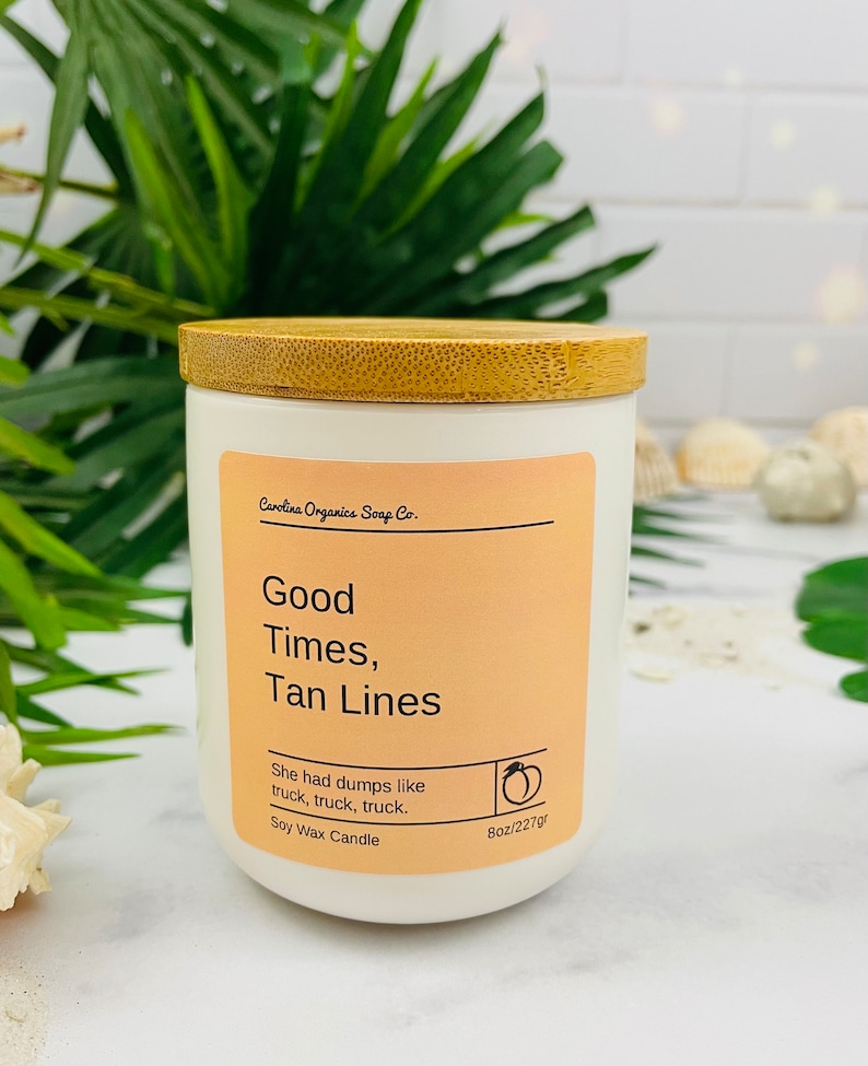 May include: A white candle with a bamboo lid. The label reads "Carolina Organics Soap Co. Good Times, Tan Lines" and "She had dumps like truck, truck, truck. Soy Wax Candle 8oz/227gr".