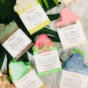 K&ouml;nnte beinhalten: Eine Auswahl handgefertigter Seifen mit verschiedenen Farben und Designs. Die Seifen sind mit Namen wie "Banana Puddin", "Cherry Blosom" und "Save the Sea Turtles" beschriftet. Die Seifen sind verpackt und mit tropischen Bl&auml;ttern und einer Muschel angeordnet.