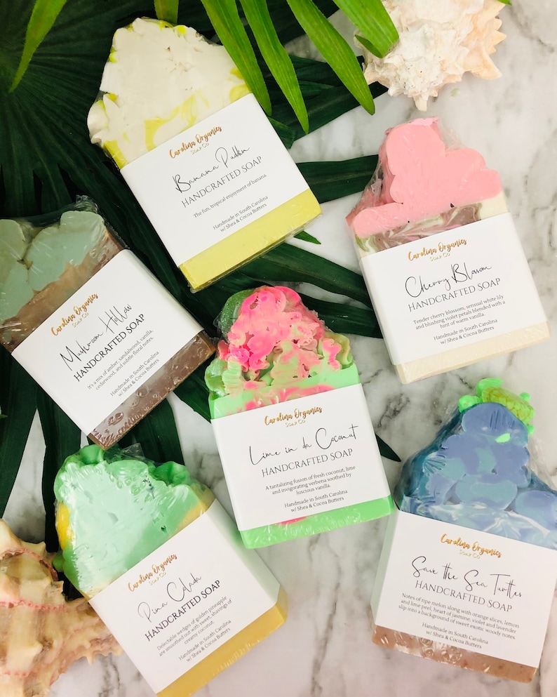 K&ouml;nnte beinhalten: Eine Auswahl handgefertigter Seifen mit verschiedenen Farben und Designs. Die Seifen sind mit Namen wie "Banana Puddin", "Cherry Blosom" und "Save the Sea Turtles" beschriftet. Die Seifen sind verpackt und mit tropischen Bl&auml;ttern und einer Muschel angeordnet.