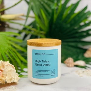 Puede incluir: Vela de cera de soja blanca con tapa de madera. La etiqueta dice "High Tides, Good Vibes" y "No shoes, no shirt, no problem." La vela pesa 227 gramos.