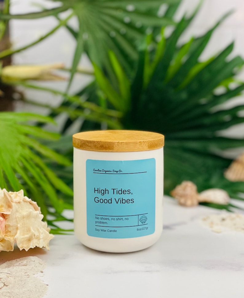 Puede incluir: Vela de cera de soja blanca con tapa de madera. La etiqueta dice "High Tides, Good Vibes" y "No shoes, no shirt, no problem." La vela pesa 227 gramos.