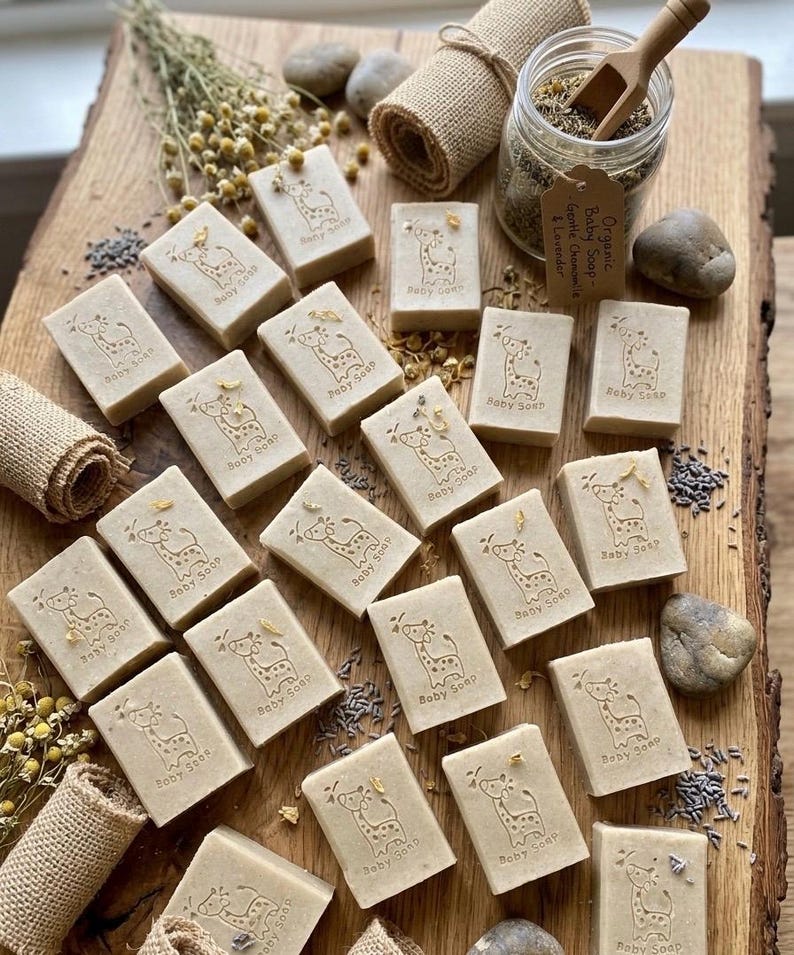 Puede incluir: Jabones rectangulares para beb&eacute;s con un dise&ntilde;o de jirafa y la inscripci&oacute;n "Baby Soap". Los jabones est&aacute;n dispuestos sobre una tabla de madera, junto con flores secas, un tarro de hierbas y peque&ntilde;as piedras. Cada jab&oacute;n mide unos 6 cm.