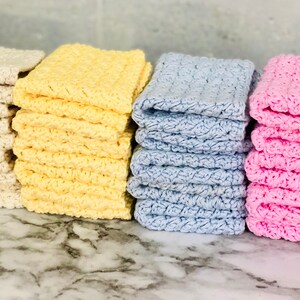 Puede incluir: Apilados de toallitas hechas a mano en varios colores: crema, amarillo, azul claro y rosa. Cada toallita tiene un dise&ntilde;o texturizado de ganchillo. Las toallitas est&aacute;n cuidadosamente apiladas, mostrando su textura suave y absorbente.