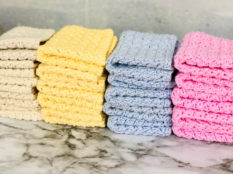 Puede incluir: Apilados de toallitas hechas a mano en varios colores: crema, amarillo, azul claro y rosa. Cada toallita tiene un dise&ntilde;o texturizado de ganchillo. Las toallitas est&aacute;n cuidadosamente apiladas, mostrando su textura suave y absorbente.