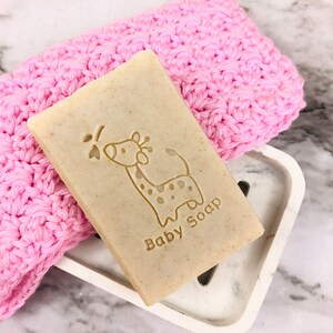 Puede incluir: Una pastilla rectangular de jab&oacute;n para beb&eacute;s con un dise&ntilde;o de jirafa y las palabras "Baby Soap" grabadas. El jab&oacute;n descansa sobre un jabonera blanca junto a un pa&ntilde;o de lavado rosa tejido. El fondo es una superficie de m&aacute;rmol.