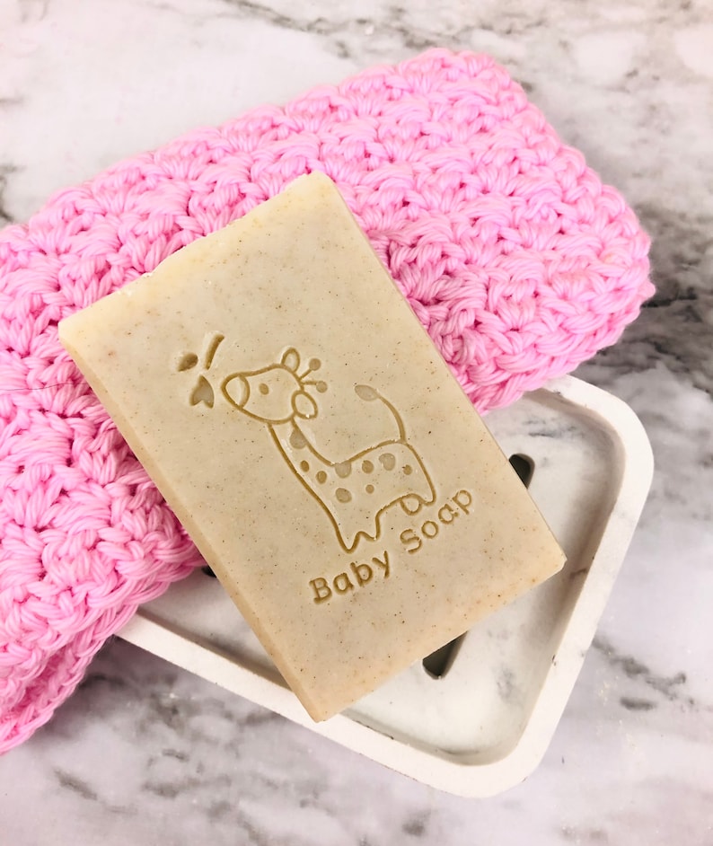 Puede incluir: Una pastilla rectangular de jab&oacute;n para beb&eacute;s con un dise&ntilde;o de jirafa y las palabras "Baby Soap" grabadas. El jab&oacute;n descansa sobre un jabonera blanca junto a un pa&ntilde;o de lavado rosa tejido. El fondo es una superficie de m&aacute;rmol.