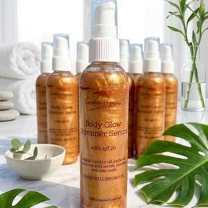 May include: Several gold-toned Body Glow Shimmer Serum bottles with white dispensers are displayed. The labels read "Body Glow Shimmer Serum with spf 20" and "Shake well before use". The background features a white towel and green foliage.