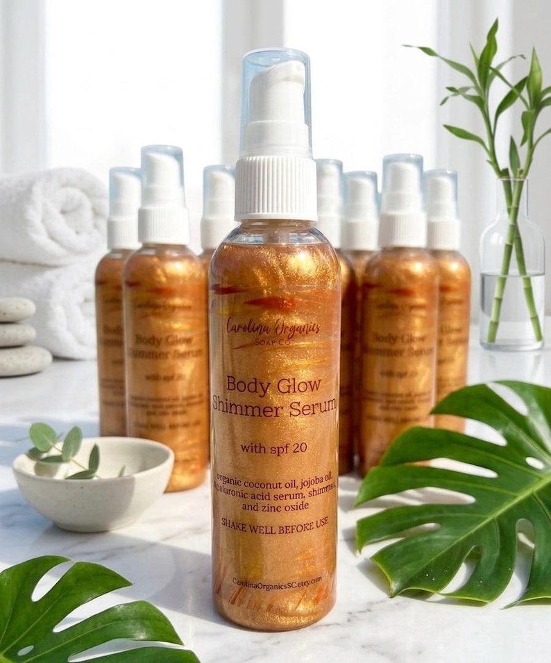 May include: Several gold-toned Body Glow Shimmer Serum bottles with white dispensers are displayed. The labels read "Body Glow Shimmer Serum with spf 20" and "Shake well before use". The background features a white towel and green foliage.
