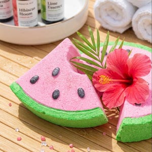 Puede incluir: Rodajas de bomba de baño con forma de sandía con centro rosa, corteza verde y detalles de semillas negras. Una flor de hibisco roja y una hoja verde se colocan en la bomba de baño. Frascos de aceites esenciales y toallas blancas enrolladas en el fondo.