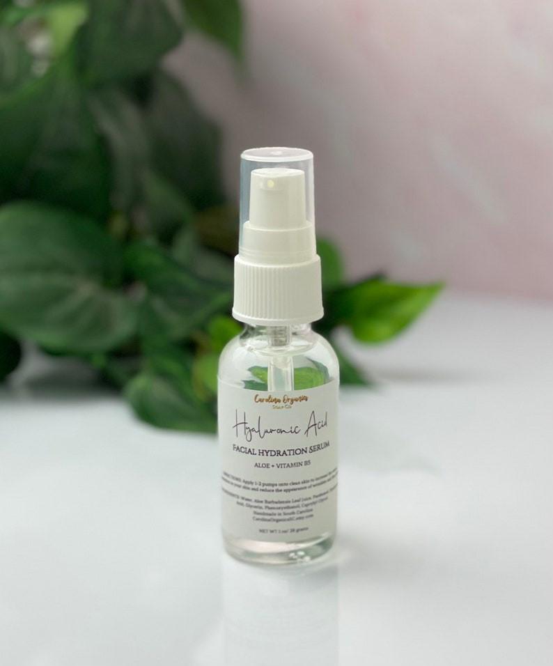 May include: A clear glass bottle with a white pump top containing Hyaluronic Acid Facial Hydration Serum. The label is white with black text and says "Aloe + Vitamin B5".
