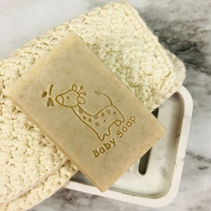 Puede incluir: Una pastilla de jab&oacute;n para beb&eacute; de color marr&oacute;n claro con un dise&ntilde;o de jirafa y la inscripci&oacute;n "Baby Soap" descansa sobre un jabonera de m&aacute;rmol blanco. Un pa&ntilde;o de lavado de ganchillo color crema est&aacute; al lado del jab&oacute;n. La jabonera tiene una ranura de drenaje.
