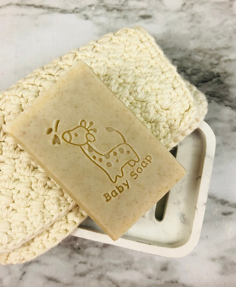 Puede incluir: Una pastilla de jab&oacute;n para beb&eacute; de color marr&oacute;n claro con un dise&ntilde;o de jirafa y la inscripci&oacute;n "Baby Soap" descansa sobre un jabonera de m&aacute;rmol blanco. Un pa&ntilde;o de lavado de ganchillo color crema est&aacute; al lado del jab&oacute;n. La jabonera tiene una ranura de drenaje.