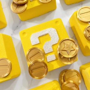 May include: Overhead shot of several yellow, question mark-adorned blocks with gold coins. The blocks have a white question mark on them. Some coins have a star in the center. The blocks are arranged on a white surface.